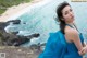 A woman in a blue dress sitting on a rock by the ocean.