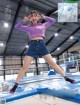 A woman in a purple shirt and blue shorts is jumping on a trampoline.